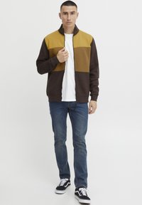 Braun und senffarbener Color-Block-Reißverschluss-Jacke, getragen über einem weißen Hemd, kombiniert mit blauen Jeans und schwarzen Turnschuhen. Baumwollmischung, lässige Passform.