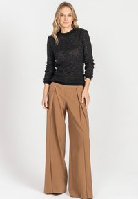 Kobieta stoi w czarnej swetrze o fakturze i brązowych spodniach o wysokim stanie i szerokich nogawkach na jasnym tle.