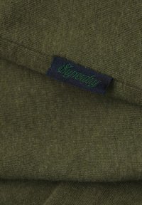 Tessuto verde oliva con una texture morbida, caratterizzato da un'etichetta blu scuro con "Superdry" ricamato in filo verde. Strati mostrati.