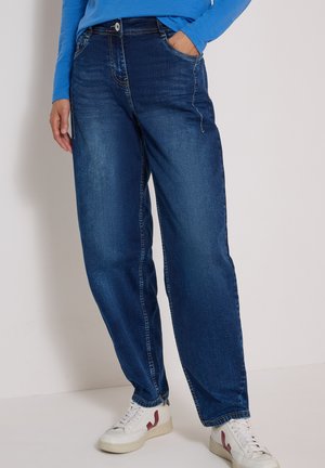 Personne portant un jean bleu foncé ample, un haut bleu à manches longues et des baskets blanches avec des touches rouges, debout contre un mur blanc.