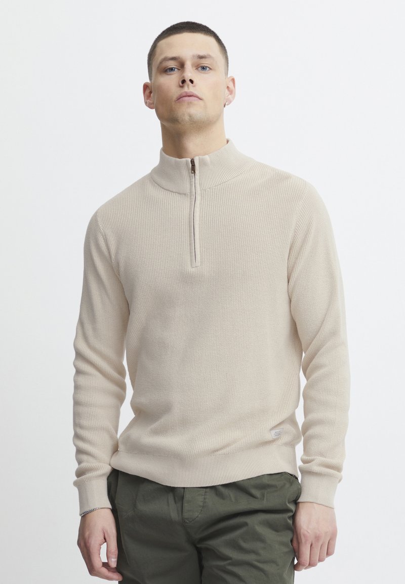 Pull en tricot côtelé beige avec un col haut et une fermeture éclair mi-longue. Il présente des manches longues et une coupe ajustée. Porté sur un pantalon vert olive.