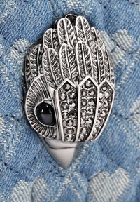 Broche en forme d'oiseau ton argent avec des détails complexes de plumes et des accents de cristal noir, posé sur un fond en denim texturé.