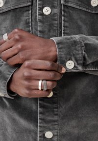 Camisa gris de botones con dos botones visibles; primer plano de manos que llevan un anillo texturizado y dos anillos de metal lisos, mostrando varios acabados.