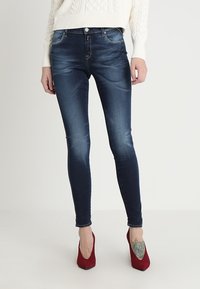 Jeans ajustados de mezclilla azul, con un acabado desgastado, bolsillos frontales y cremallera. Se llevan con zapatos de tacón altos rojos y un suéter blanco.
