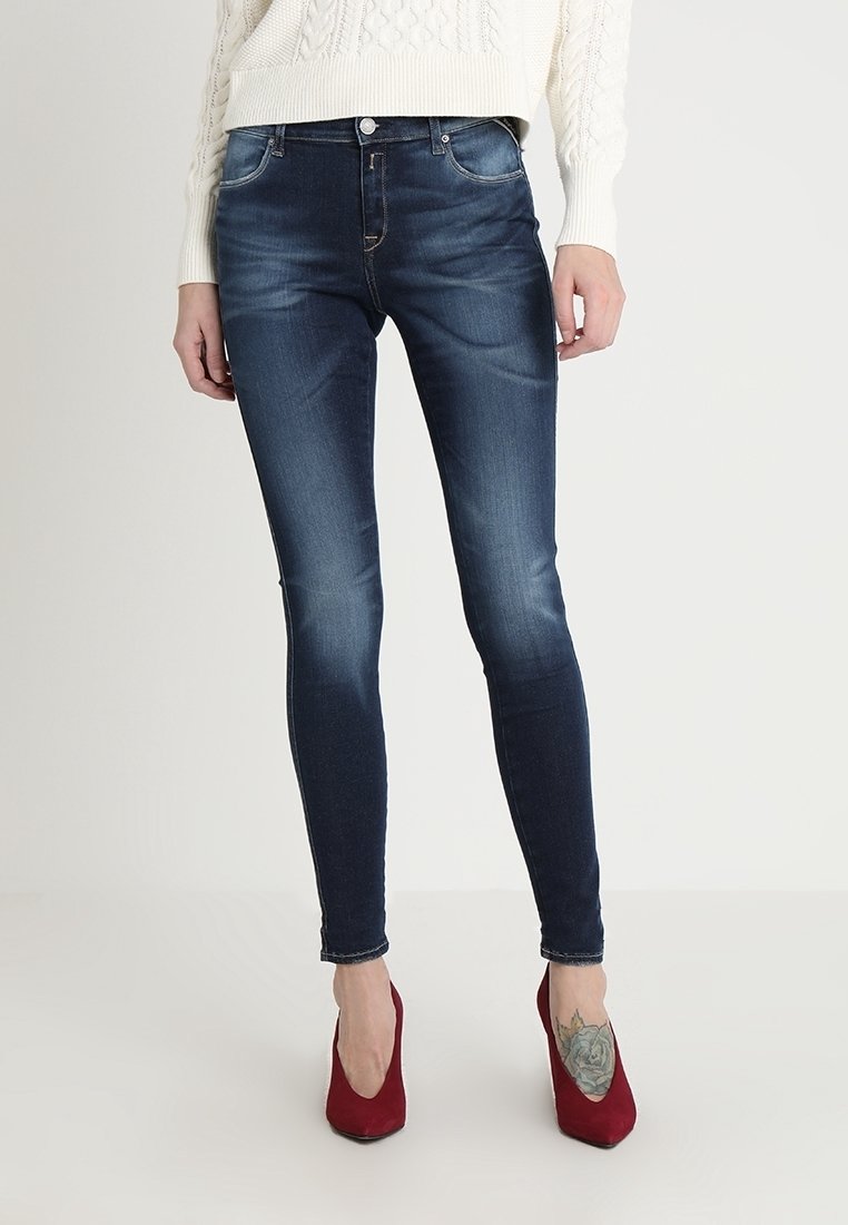 Jeans ajustados de mezclilla azul, con un acabado desgastado, bolsillos frontales y cremallera. Se llevan con zapatos de tacón altos rojos y un suéter blanco.