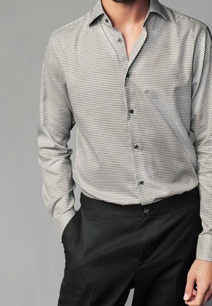 Homme portant une chemise à manches longues à petits carreaux noir et blanc, rentrée dans un pantalon habillé noir, une main dans la poche.