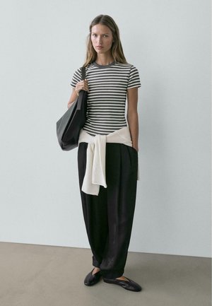 Kobieta ubrana w szarą i białą koszulkę w paski, czarne spodnie o szerokich nogawkach, czarne buty, biały sweter zawiązany wokół talii oraz niosąca czarną torbę.