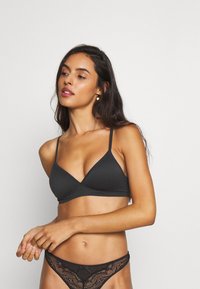 Zwarte driehoekige bralette met dunne verstelbare bandjes. Zacht materiaal met een gladde textuur en minimale stikdetails. Gecombineerd met een thonga met kant uitsnijdingen.