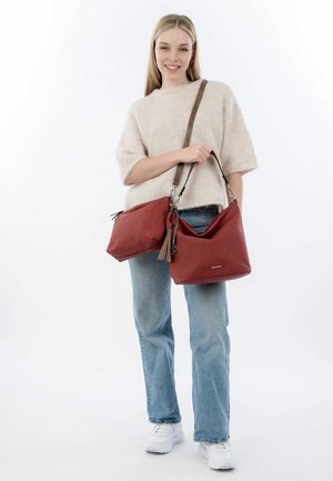 Sac à main en cuir rouge au design décontracté, avec une bandoulière amovible en cuir marron et des détails en franges, posé sur un fond blanc.