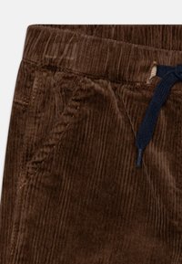 Pantalones de pana marrón con cintura ajustable mediante cordón. La tela tiene costillas verticales, y la cinturilla cuenta con un lazo azul marino para el ajuste.