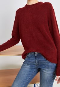Personne portant un pull en maille rouge et un jean bleu, appuyant le bras gauche sur une surface en bois devant un fond uni.