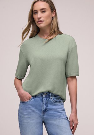 Mujer con cabello castaño claro, vestida con un top de manga corta de punto verde claro y jeans azul claro, de pie con una mano en el bolsillo contra un fondo sencillo.