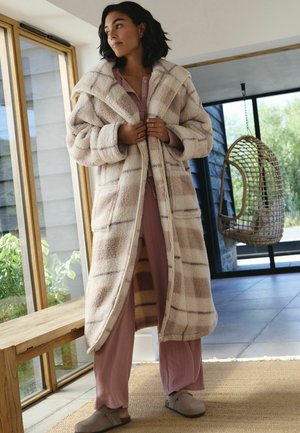 Flauschiger beiger und cremfarbener karierter langer Mantel mit großen Revers, Fronttaschen und einem vollständigen Reißverschluss, kombiniert mit hellrosa Loungewear und Hausschuhen.