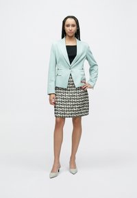 Gaiši zils piegriezts žakete ar diviem pogām, valkāts pār melnu topu, kombinēts ar melnu un baltu rakstu svārku un pelēkiem augstpapēžu apaviem.