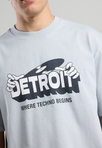 Light gray t-shirt featuring a bold black graphic with the word "DETROIT" and text "WHERE TECHNO BEGINS," printed on smooth fabric.