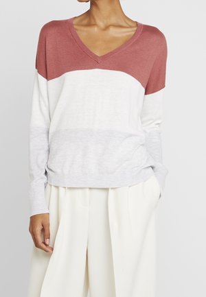 Woman wearing a color-block V-neck sweater with dusty rose, white, and light gray sections, paired with loose white pants.