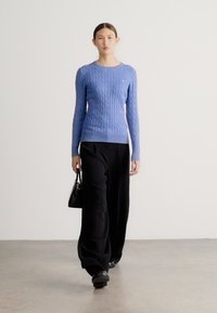 Zilā džempera ar rakstu pinumu, ar pieguļošu formu, kombinēts ar vaļīgām melnām biksēm un mazu melnu somiņu. Gluda tekstūra un vienkārša silueta.