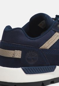 Marinblå sneakers med texturerad mocka, beige detaljer och mörkblå sömmar. Har en logotyp och en kontrasterande vit mellansula med mönster.