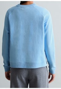 Personne portant une chemise bleu clair à manches longues et un pantalon gris, vue de dos sur un fond clair uni.