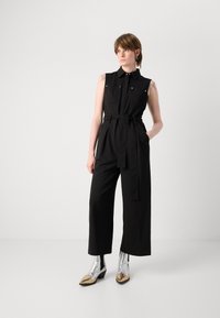 Mono negro sin mangas con dos bolsillos en el pecho, cinturón en la cintura y piernas palazzo, combinado con botas plateadas hasta el tobillo y punta dorada.