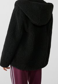 Schwarze Fleece-Jacke mit Kapuze, die eine strukturierte, flauschige Oberfläche und eine lockere Passform aufweist. Sichtbare Nähte entlang der Nähte und Ränder.