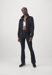 Kvinde iført sort sportjakke, sorte flare leggings, hvid sports-bh og sorte løbesko, stående mod en ensfarvet baggrund.