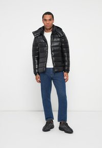 Giacca a vento nera con finitura lucida, chiusura con zip e cappuccio, abbinata a una t-shirt bianca e jeans blu. Tessuto testurizzato, design trapuntato.
