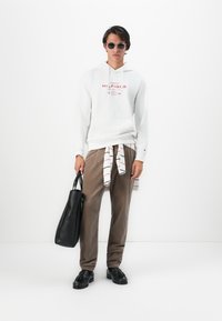 Jeune homme portant des lunettes de soleil, un sweat à capuche blanc Tommy Hilfiger, un pantalon marron, des mocassins noirs, tenant un sac fourre-tout noir, avec une chemise rayée nouée autour de la taille.