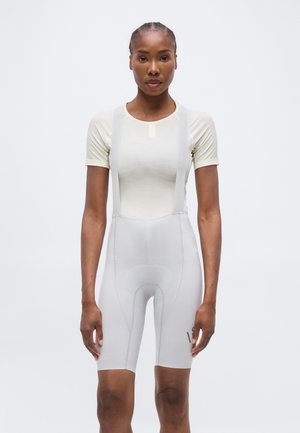 Kobieta z zaplecionymi włosami, ubrana w dopasowaną krótki rękaw bluzkę oraz jasne rowerowe spodenki na szelkach, stojąca na jednolitym tle.