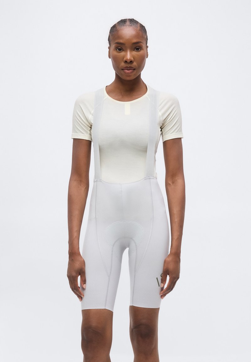 Γυναίκα με πλεξούδες, ντυμένη με εφαρμοστή κοντομάνικη μπλούζα και ανοιχτόχρωμα ποδηλατικά σορτς με τιράντες, στέκεται μπροστά σε απλό φόντο.