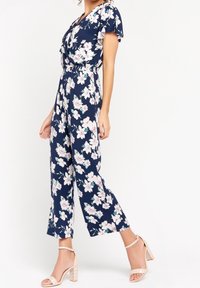 Námořnický květinový overal s krátkými volánovými rukávy, elastickým pasem a širokými zkrácenými nohavicemi; má vzory bílých a růžových květin.