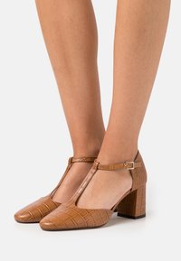 Brune faux læder T-strop hæle med krokodillemønster, firkantet tå og blokhæle; har en sidelukning.