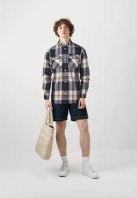 Camisa de franela a cuadros con patrones en azul, blanco y marrón; dos bolsillos en el pecho y cierre de botones. El modelo también lleva pantalones cortos de navy y sostiene una bolsa beige.