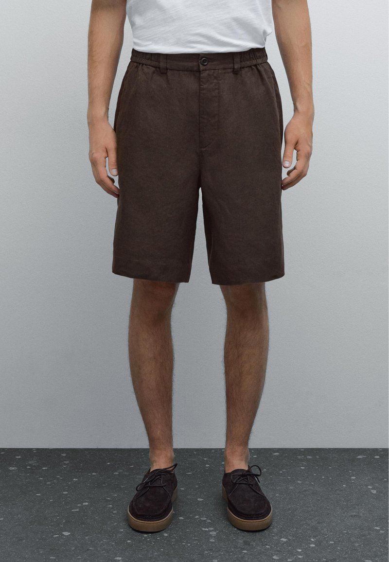 Shorts en lin marron avec une coupe décontractée, taille élastique et poches avant, associés à des chaussures en daim foncé sur une surface texturée.