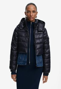 Giacca nera imbottita con cappuccio removibile, dotata di zip sul davanti e dettagli in denim nella parte inferiore. Tessuto testurizzato con design trapuntato.