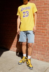 Gul Lakers-t-shirt med lila bokstäver, jeansshorts med fransig fåll och svarta/gula höga sneakers. Tegelvägg i bakgrunden.