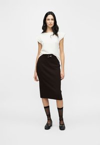 Λευκό τοπ με κοντά μανίκια, ριμπ καφέ μίντι φούστα με θηλιές ζώνης και μεταλλική λεπτομέρεια. Το μοντέλο φοράει διάφανα μαύρα καλσόν μέχρι το γόνατο και μαύρα παπούτσια.