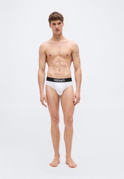 Jeune homme debout, pieds nus, portant un slip blanc avec une ceinture noire affichant le nom de la marque Versace.