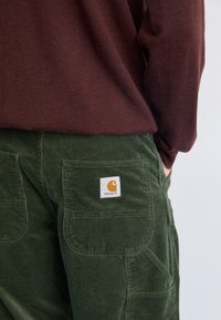 Pantalones de pana verde oscuro con un acabado texturizado, que cuentan con bolsillos traseros y un parche de logo blanco de Carhartt en el bolsillo derecho.