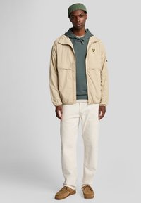 Beige lichtgewicht jack met een capuchon, voorzakken, gedragen over een groene hoodie, witte broek en bruin moccasinschoenen. Olijfgroene beanie op het hoofd.