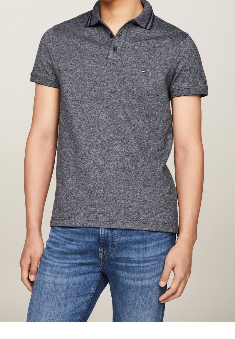 Homme portant un polo gris chiné à manches courtes avec un petit logo sur la poitrine et un jean bleu, debout devant un fond beige uni.