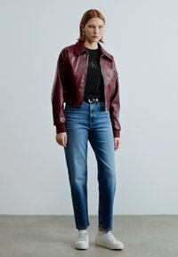 Μπουφάν από δερμάτινο μπορντό με γιακά πάνω από μαύρο T-shirt, συνδυασμένο με τζιν ελαφρύ μπλε με ίσιο καβάλο και λευκά αθλητικά παπούτσια. Λεία υφή.