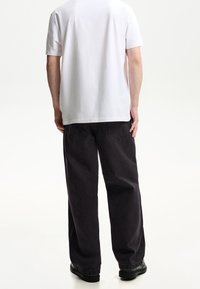 En person som bär en enkel vit t-shirt med korta ärmar, lösa svarta jeans och svarta skor, stående mot en vit bakgrund.