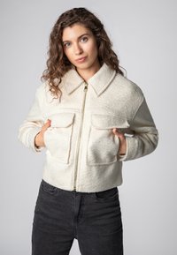 White, textured jacket with a collar, front zipper, and two chest pockets. Styled with black pants, against a gray background.
