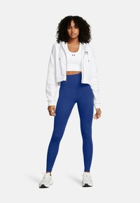 Donna con capelli ricci che indossa un hoodie cropped bianco, un reggiseno sportivo bianco, leggings a vita alta blu e sneakers bianche, in piedi su uno sfondo bianco.
