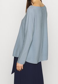 Světle modrý svetr s volným, širokým střihem; strukturovaná látka; dlouhé rukávy; obsahuje nenápadný detail knoflíku na zádech. Kombinováno s tmavou sukní.