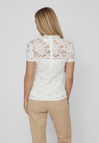 Top bianco in pizzo con motivi floreali, maniche corte, scollo a scallop e chiusura a occhio di pernice sulla schiena. Abbinato a pantaloni beige.