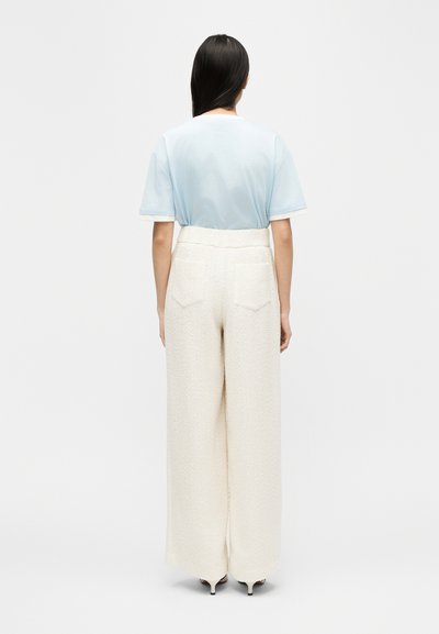 Γυναίκα με μακριά σκούρα μαλλιά, φοράει γαλάζιο τοπ με κοντά μανίκια και ψηλόμεσο λευκό παντελόνι με φαρδιά μπατζάκια και υφή, στέκεται με την πλάτη γυρισμένη.