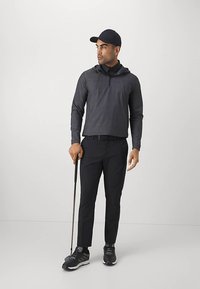 Vyrų tamsiai pilkas kapučino puloveris su pusiau užtrauktuku, derinamas su juodomis kelnėmis ir juodais golfo batais. Laiko golfo lazda.