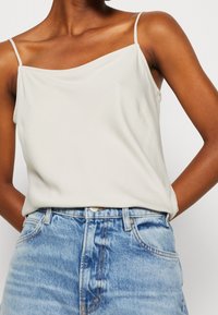 Top in off-white, in tessuto leggero con una texture liscia, dotato di spalline sottili e una vestibilità comoda. Abbinato a jeans blu a vita alta.